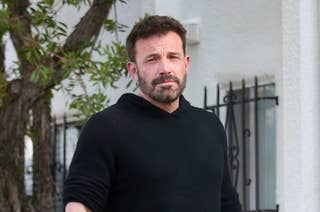 Ben Affleck is outdoors, wearing a casual black hoodie. There's a tree and a white building with bars on the windows in the background