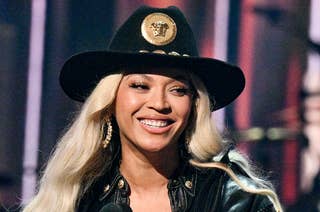 Beyoncé smiles while holding a trophy and speaking into a microphone on stage. She wears a black fringed leather jacket and a black hat