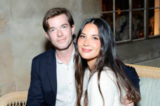 Olivia Munn and John Mulaney smiling, sitting close together on a bench, Olivia in a sweater and jeans, John in a blazer and shirt