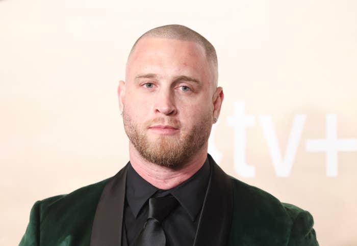 Chet Hanks with a shaved head and beard wearing a velvet suit jacket and black shirt stands in front of a backdrop with the Apple TV+ logo