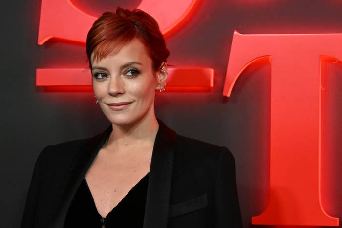 Lily Allen stands against a dark background with neon red letters, wearing a black blazer over a black top