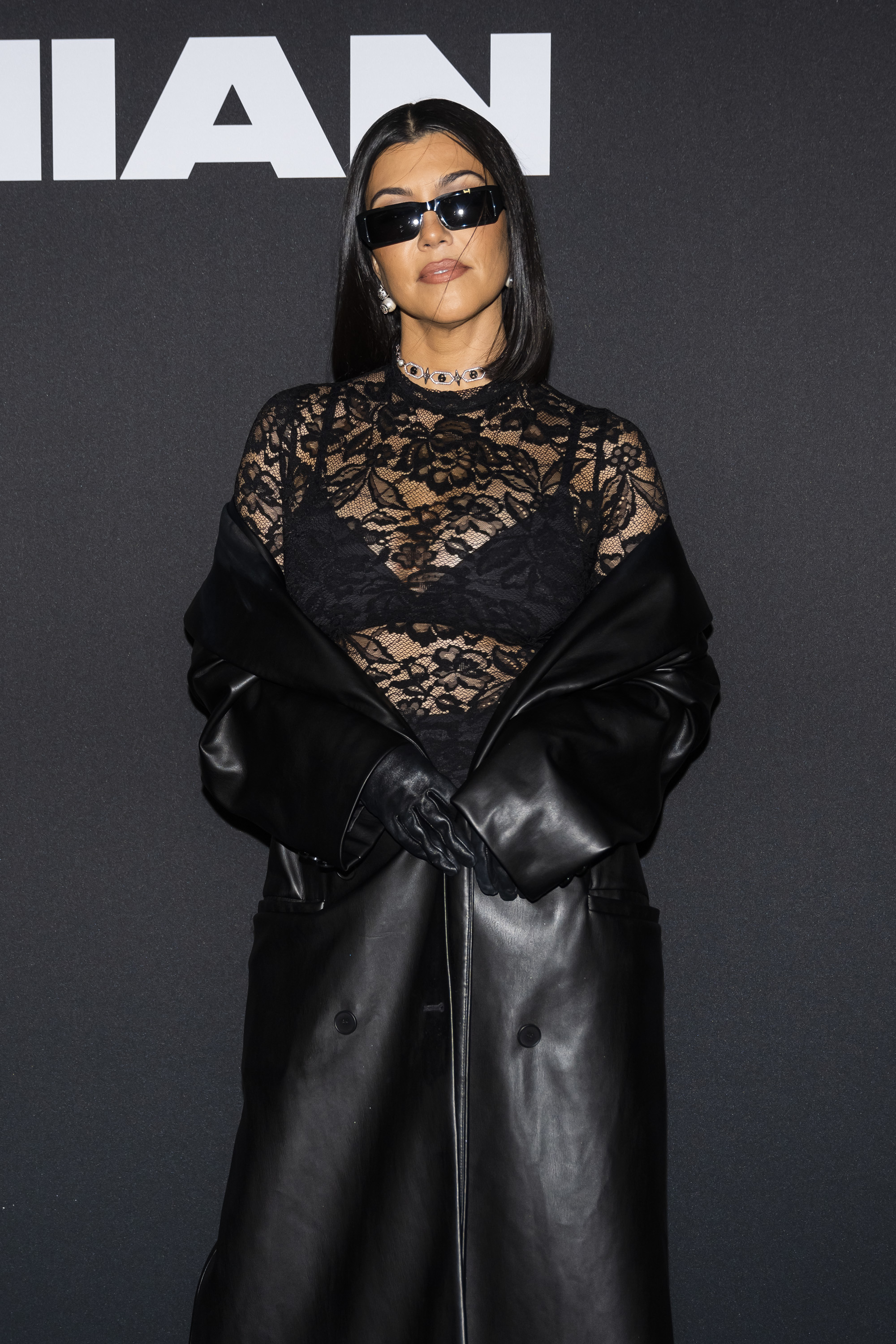 Kourtney Kardashian at an event in a lace top with black bra underneath, leather gloves, and long leather coat, posing against a dark background