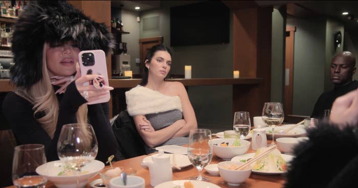 Khloé Kardashian takes a selfie while Kendall Jenner, in a stylish off-shoulder dress, and others sit at a dinner table with various dishes