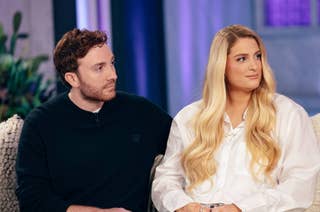 Daryl Sabara and Meghan Trainor sitting on a couch during a television interview. Daryl is in casual attire, and Meghan is wearing a white blouse with relaxed waves in her hair