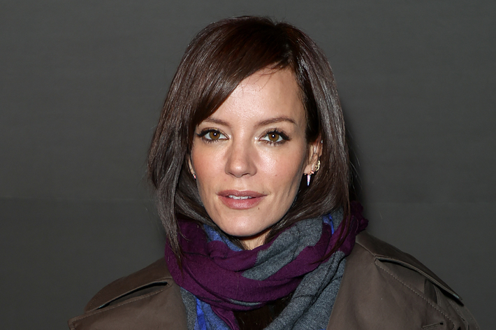 Lily Allen wearing a dark coat and a colorful scarf, holding a white fluffy bag. She has short, styled hair and is posing for the camera