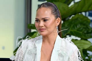 Chrissy Teigen in a light, feathery jacket with pulled-back hair, wearing drop earrings, in front of green plants