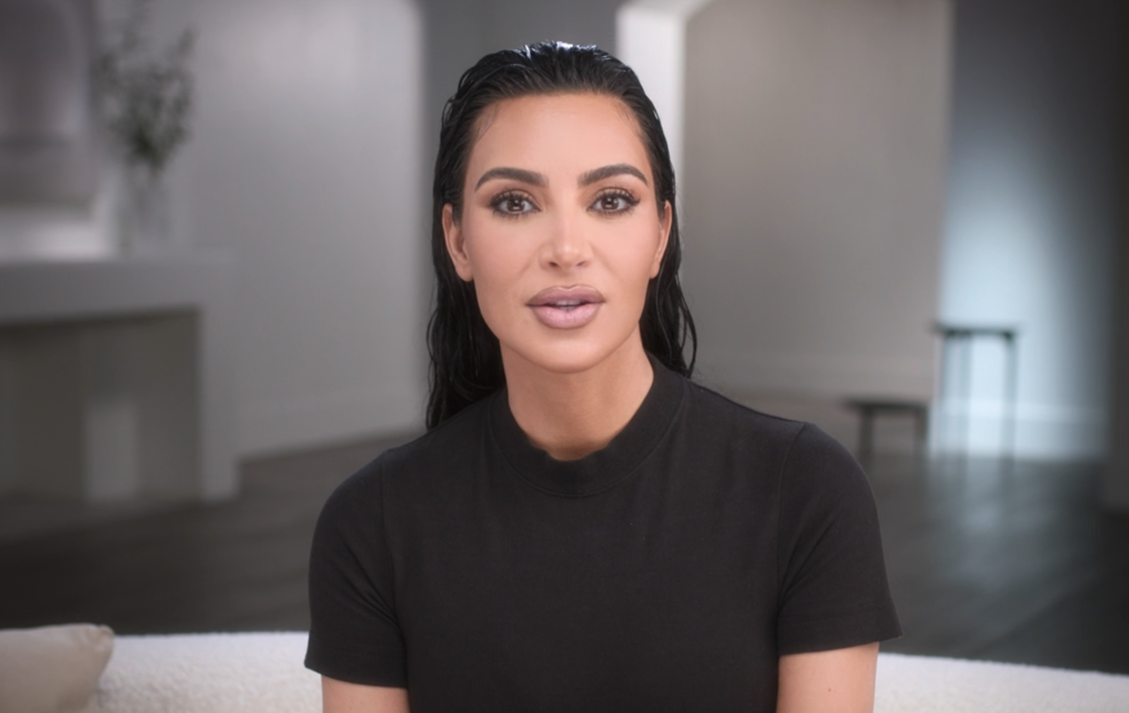 Kim Kardashian sits on a couch in a minimalistic room, wearing a simple black top, with slicked-back hair and natural makeup