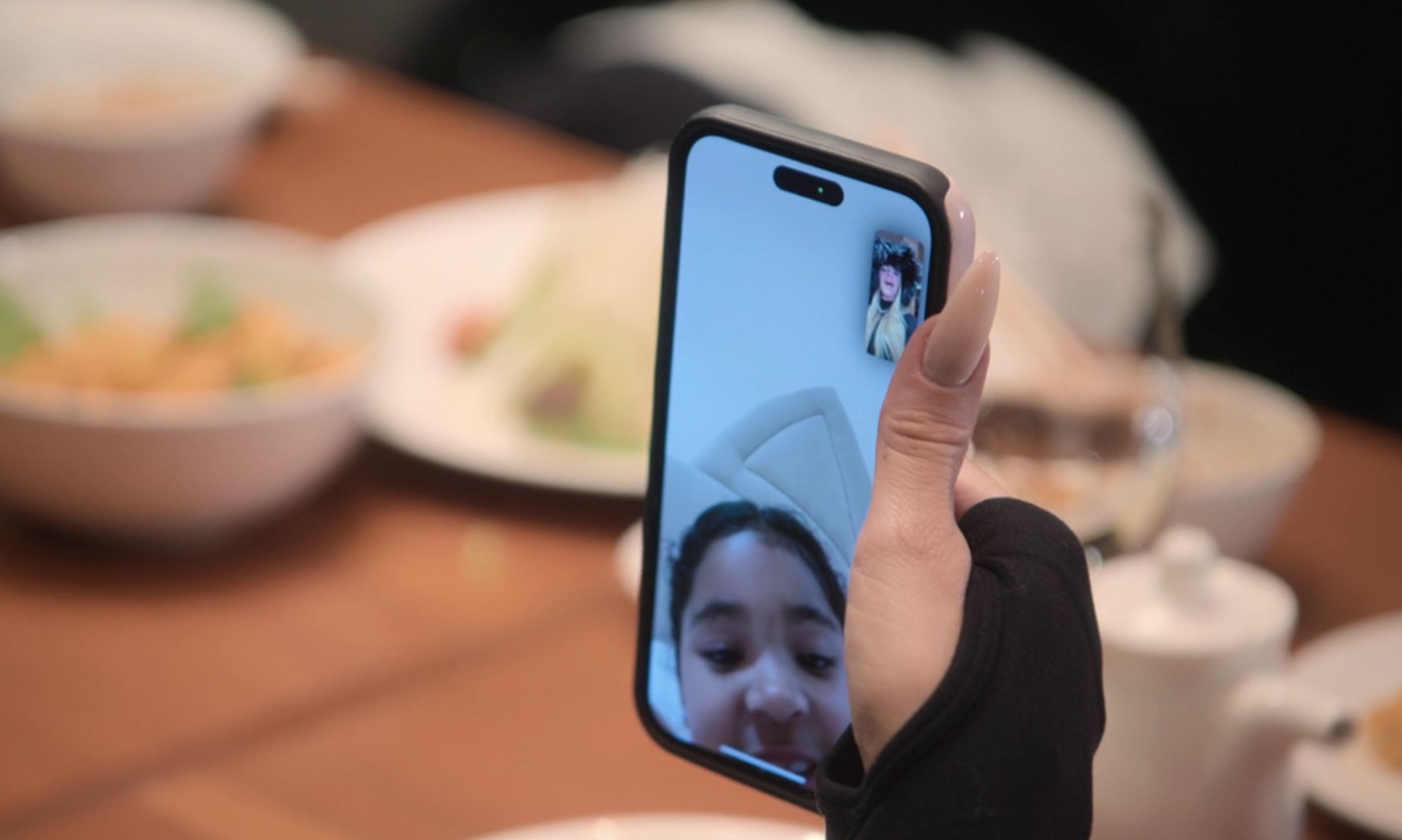 Close-up of a hand holding a phone showing a video call with two people. In the background, a dining table with several plates of food is visible