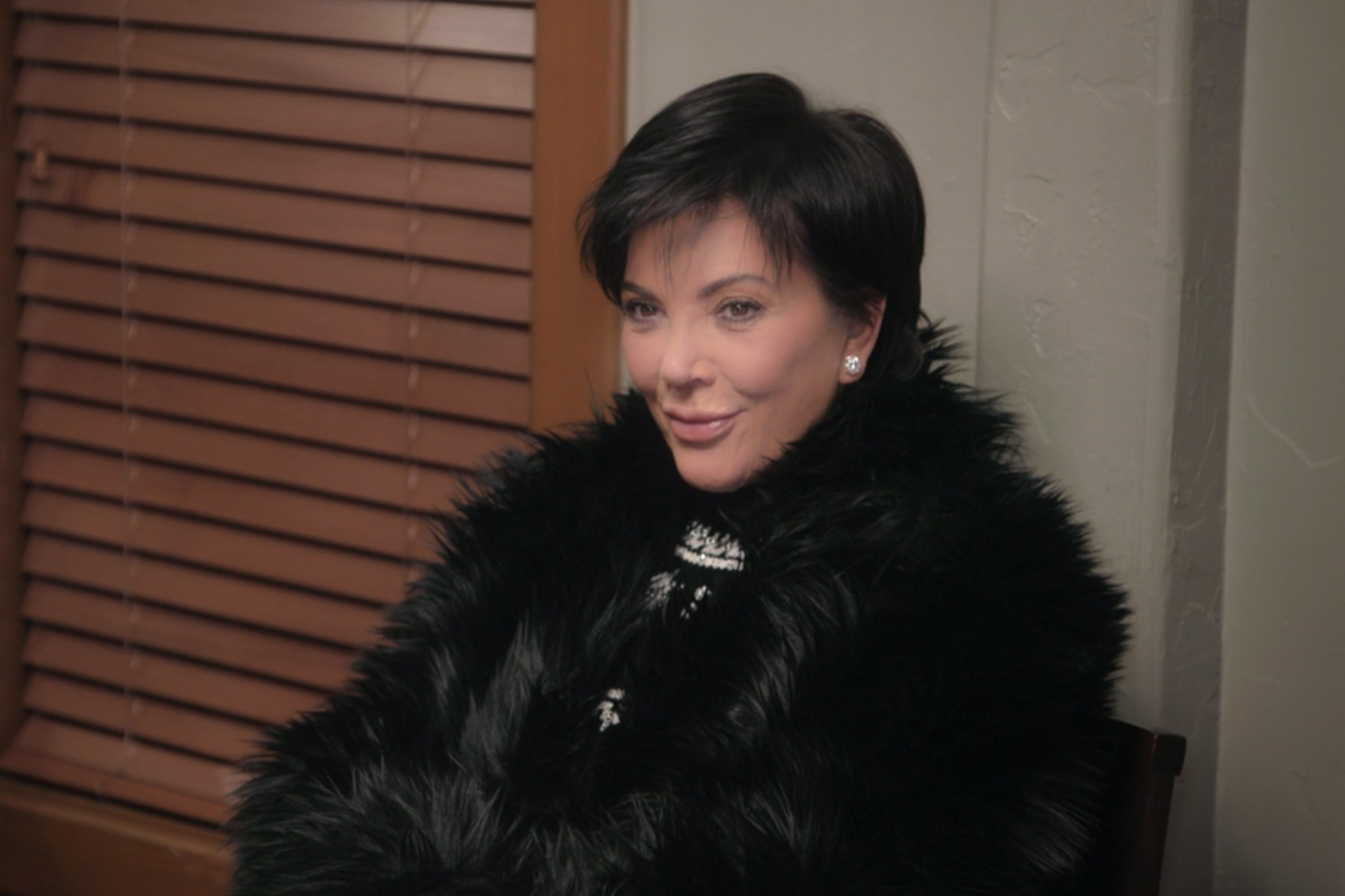 Kris Jenner is seated indoors, wearing a black fur coat with a patterned top underneath. She is smiling and looking slightly off-camera