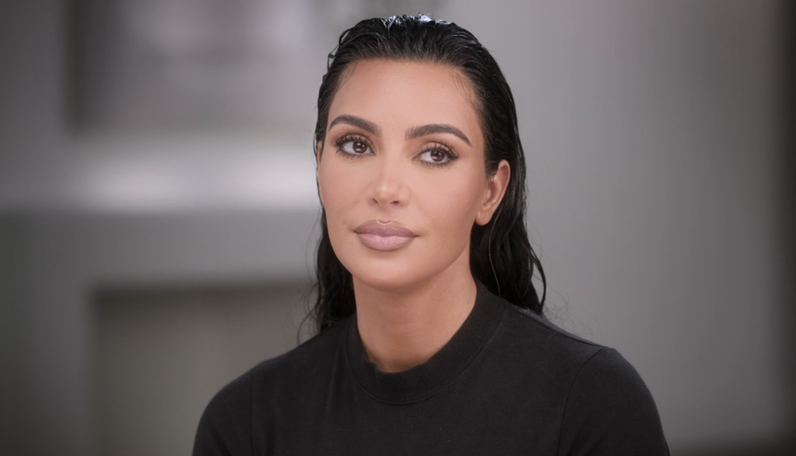 Kim Kardashian, hair slicked back, wearing a fitted black top, appearing in an interview setting