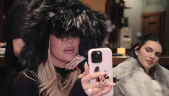 Khloé Kardashian is taking a selfie while wearing a large, fluffy hat. Kendall Jenner sits behind her, also wearing a fur coat