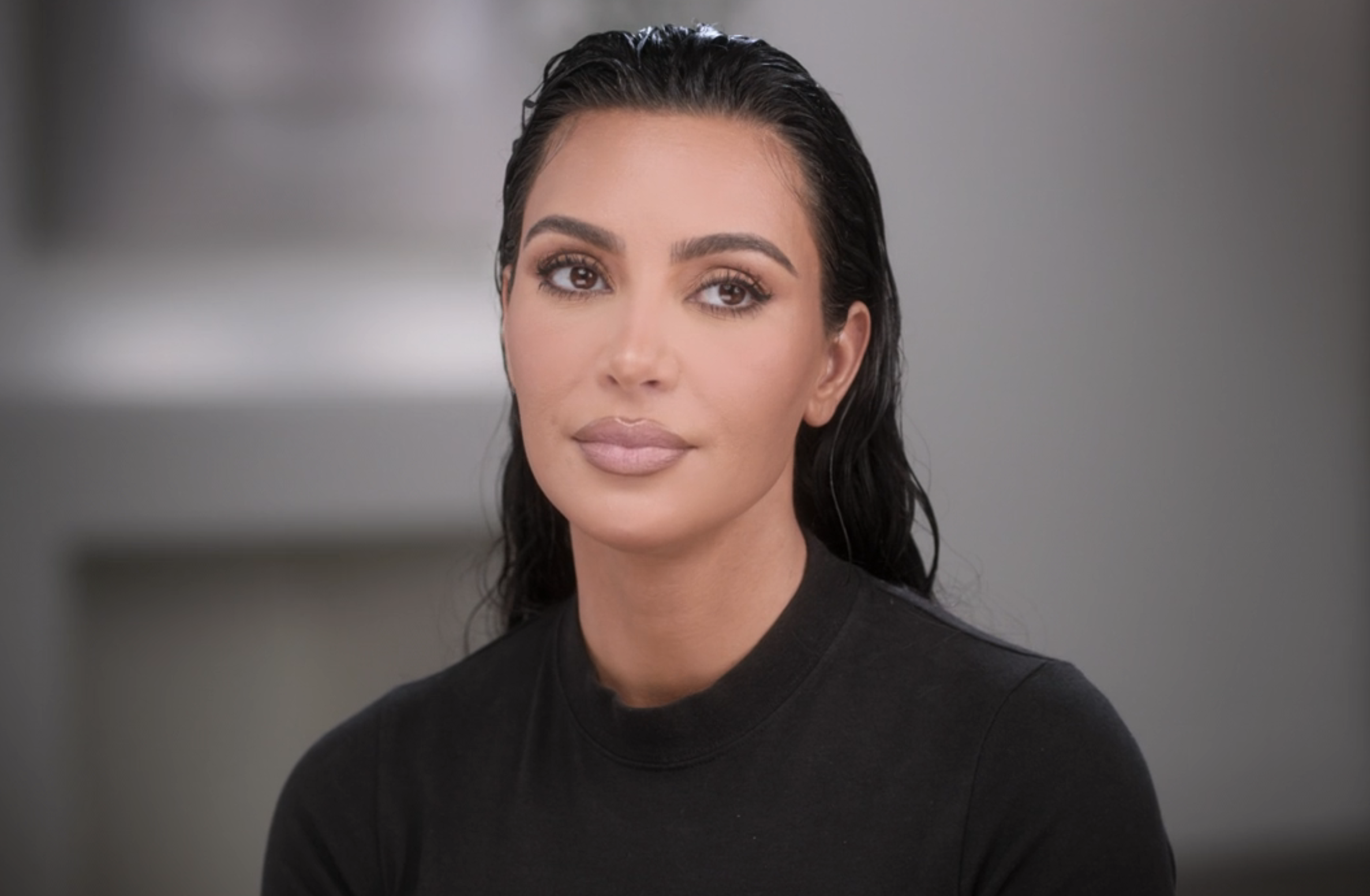 Kim Kardashian, with slicked-back hair, is wearing a simple, fitted black top. She has a calm, neutral expression and is looking slightly off-camera