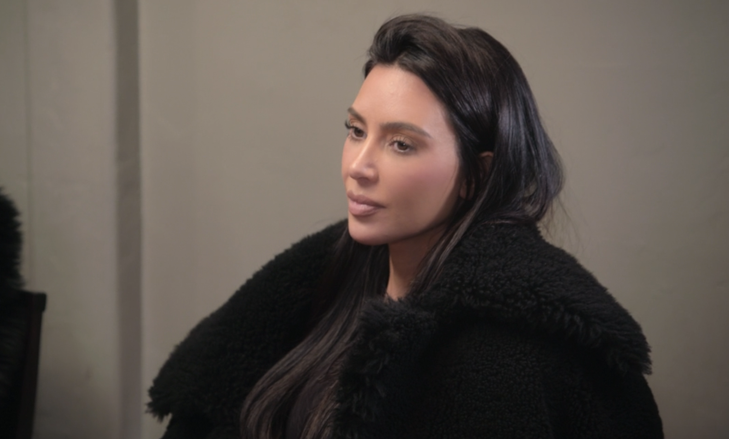 Kim Kardashian sits in a formal setting, wearing a black fur coat