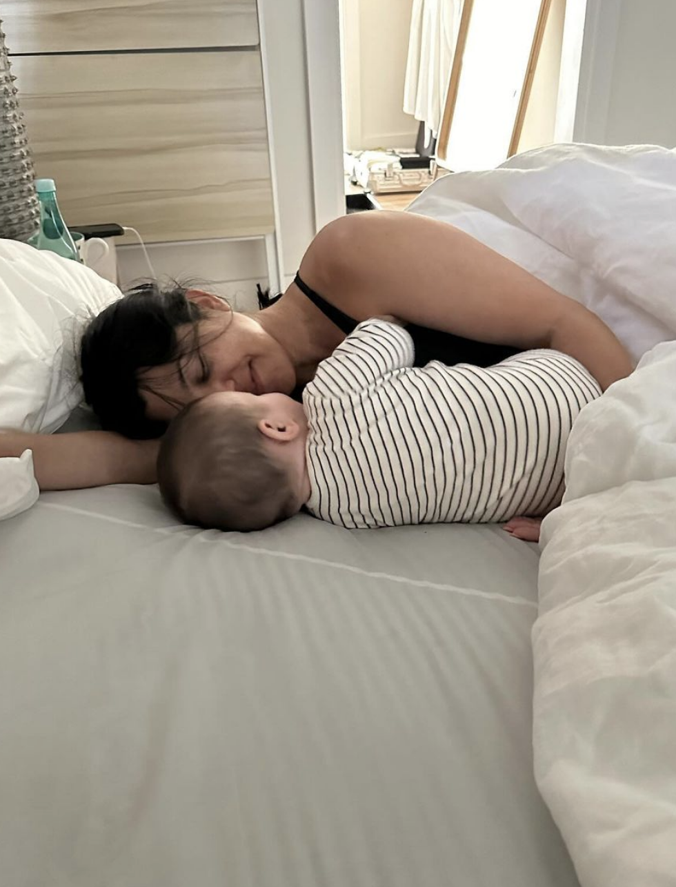 Kourtney Kardashian lies in bed facing a baby in striped clothing, nose to nose, while both rest on their sides under a white blanket