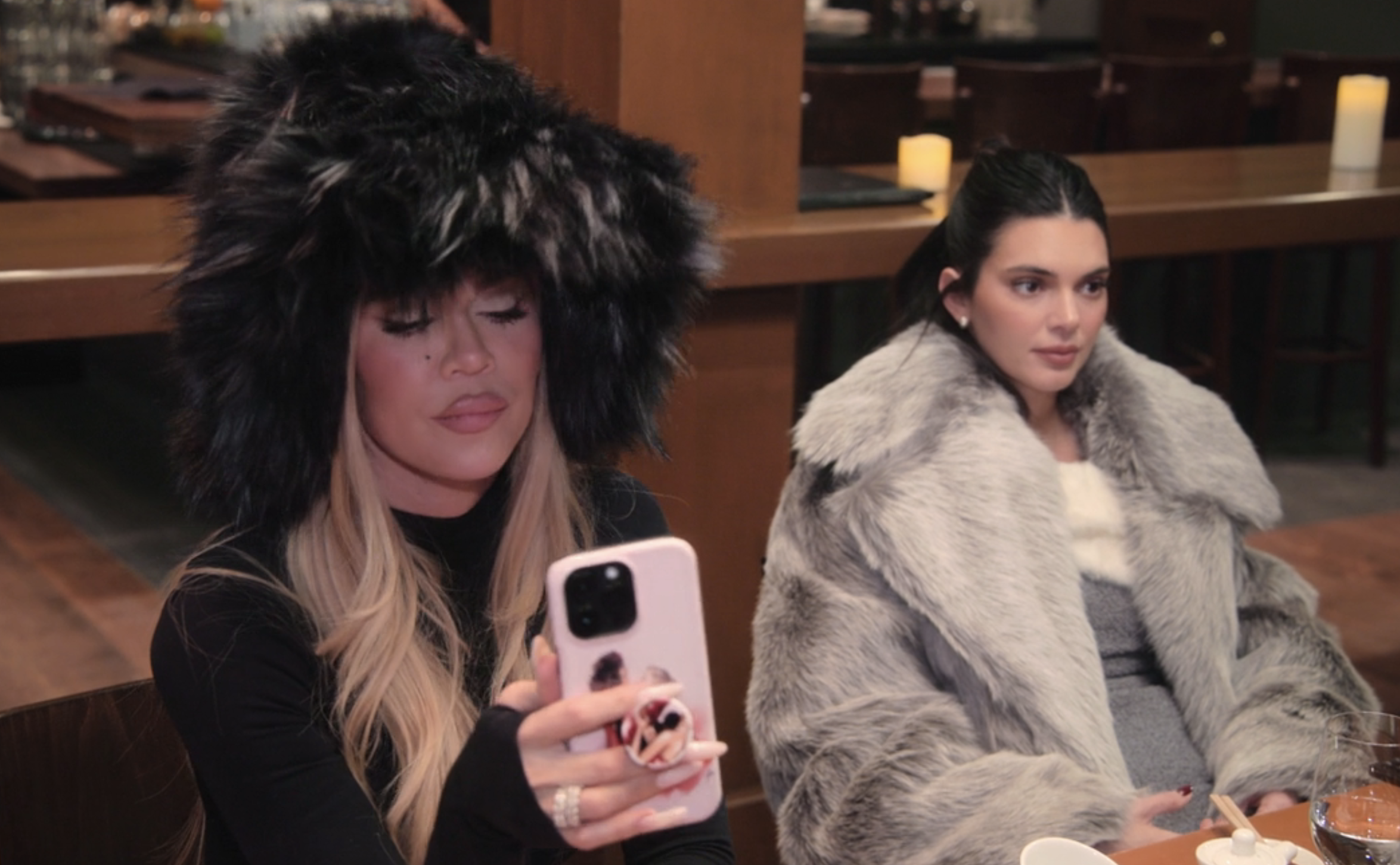 Khloe Kardashian and Kendall Jenner sit at a table; Khloe scrolls on her phone wearing a large faux-fur hat, while Kendall wears a gray faux-fur coat