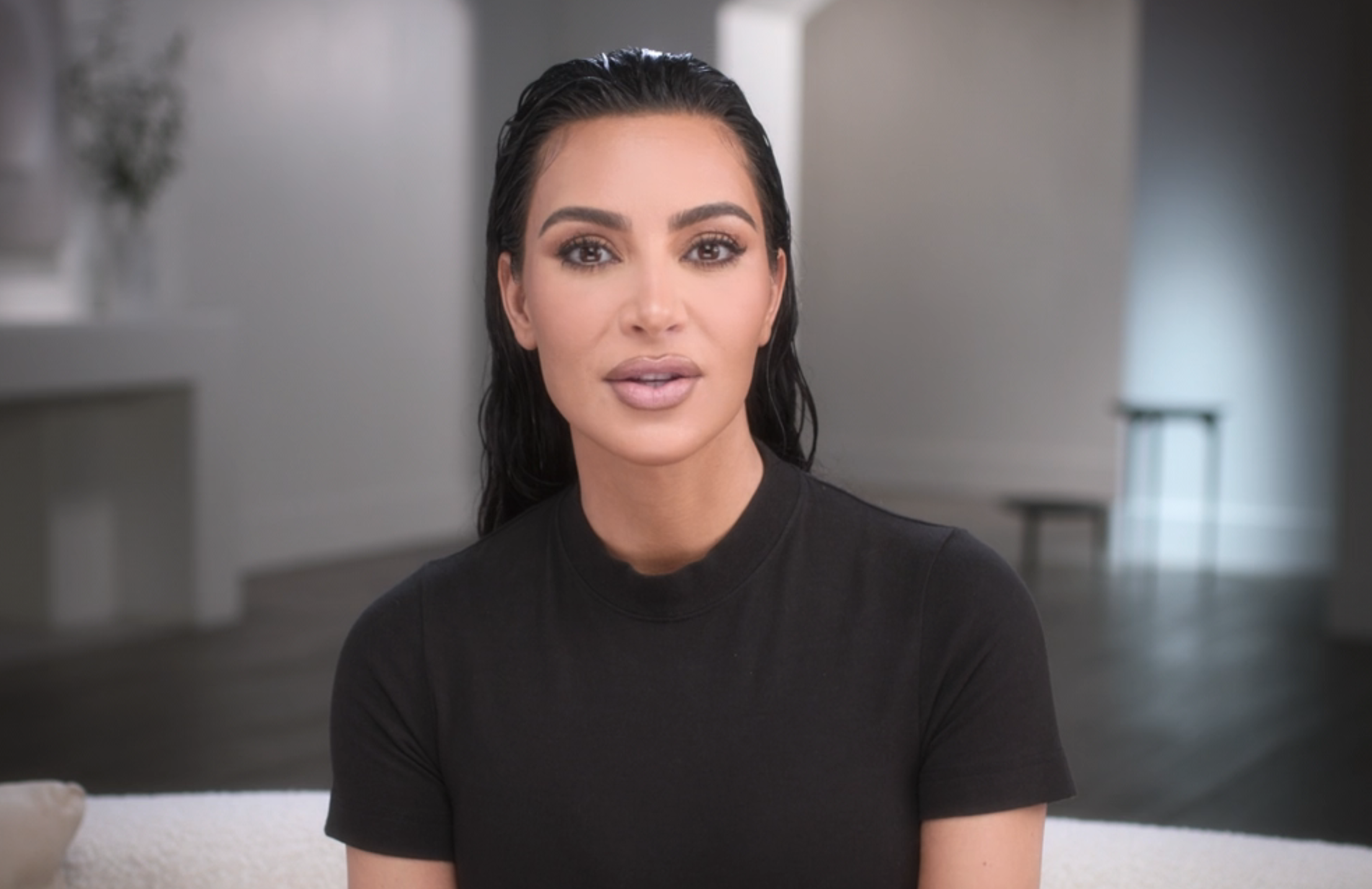 Kim Kardashian, with wet hair and wearing a simple black shirt, sits on a light-colored couch in a modern, minimalistic room